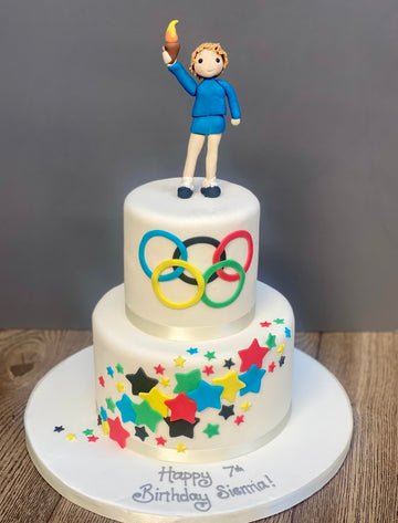 Two-tiered birthday cake with Olympic rings and star decorations, topped with a figurine of a person holding a torch.