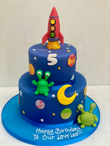 Two-tiered birthday cake with space-themed decorations including a rocket, planets, and aliens