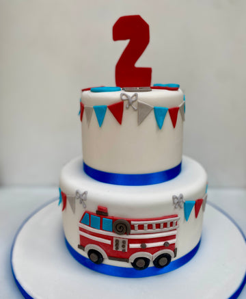 Two-tier birthday cake with fire truck design and number '2' on top, against a white background with bunting