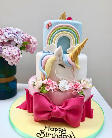 3 tier unicorn cake decorated with a unicorn on the front of the cake, a rainbow , sugar flowers and a large pink bow