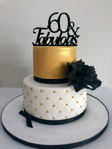Two-tiered cake with gold and white design, black floral decoration, and '60 & Fabulous' topper.