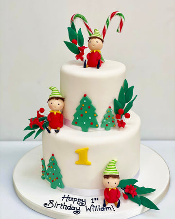 Two-tiered birthday cake with Christmas-themed decorations, including elves and candy canes,