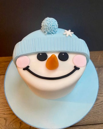 Snowman-themed Christmas cake with a blue hat and carrot nose on a wooden surface