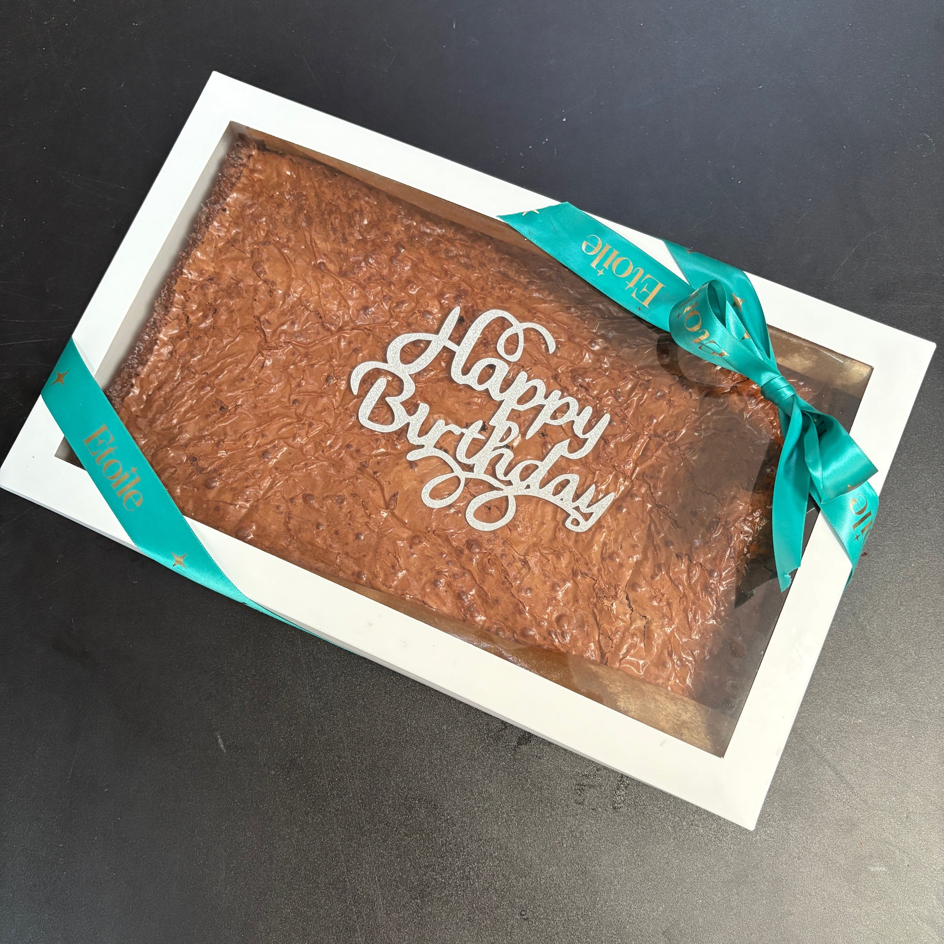 Brownie in a box with 'Happy Birthday' text and teal ribbon on a dark background