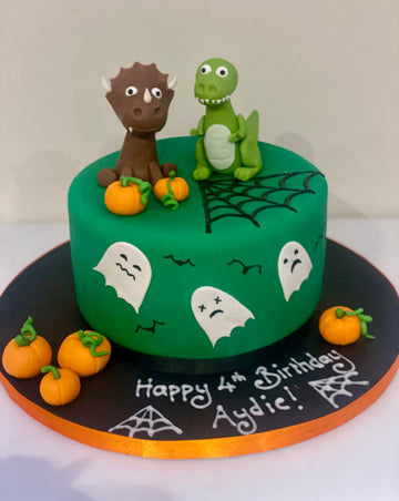 Green birthday cake with dinosaur figures, pumpkins, and Halloween-themed decorations on a white background.