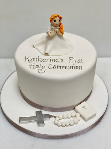 White cake with 'Katherine's First Holy Communion' text, little girl praying angel figurine, cross, and rosary on a white background