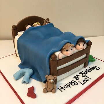 Children's birthday cake with a bed scene, wtih lots of boys playing in the bed, a teddy bear, and 'cloths scattered about