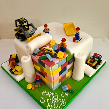 Birthday cake with lego toy construction vehicles and figures on and logo pieces showing on the side of the cake