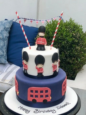 Two-tiered birthday cake with London-themed decorations including a bus and British flags, on a table outdoors.