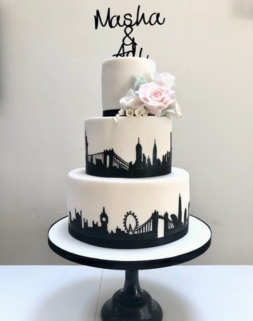 3 Tier wedding cake with the silhouette of london's skyline on the bottom tier, new york's skyline in silhouette on the middle tier - both hand painted. Top tier has a cake topper and pink sugar roses and blossoms