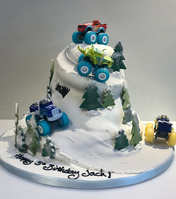 Birthday cake with toy monster truck cars on a snowy mountain landscape