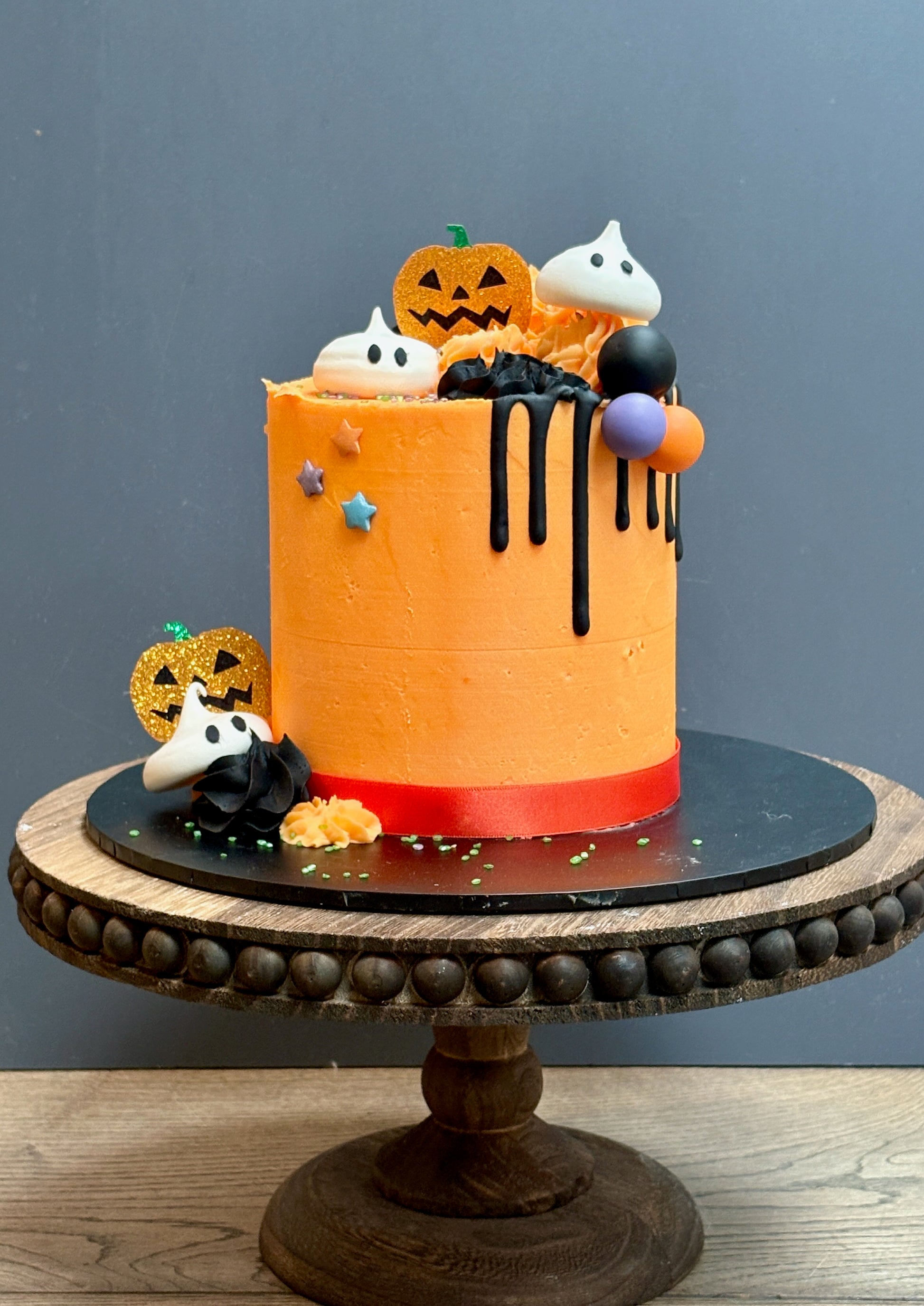 Halloween themed cake with meringue ghost decorations, a black drip, sparkly pumpkins and halloween balls