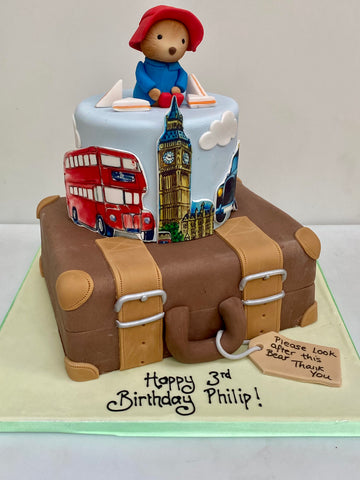 Paddington bear sat on a birthday cake to look like london and a suitcase