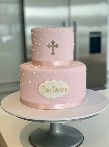 Two-tiered pink communion cake with a cross and 'Ella Quinn' text on a white surface.