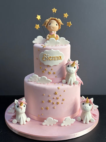 Pink two tier cake with three unicorns by the side and a little sugar girl on top