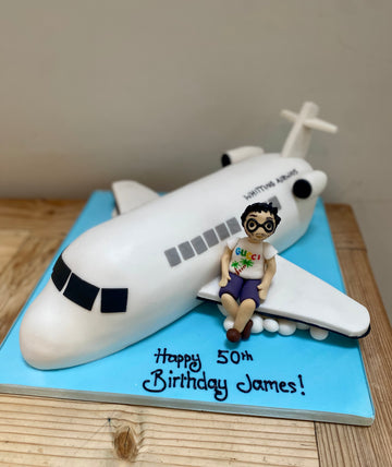 Plane-shaped birthday cake with a figurine on top, featuring 'Happy 50th Birthday James!' text.