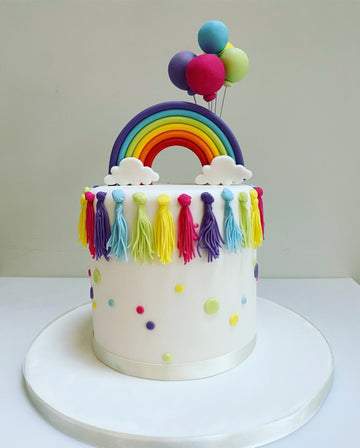 Tall white birthday cake with rainbow tassles down the sides, rainbow on top and rainbow balloons