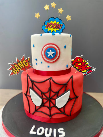 Two-tiered cake with Spider-Man and Captain America designs, featuring 'BOOM', 'BANG', and 'POW' toppers.