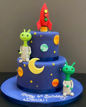 Two-tiered birthday cake with space-themed decorations including a rocket, alien, moon, and planets on a dark background.