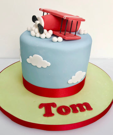 Cake with a red airplane on top, blue sky background, and 'Tom' text.