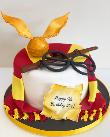 Single tier birthday cake with decorations of snitch, glasses, wand, and hogwarts scarf