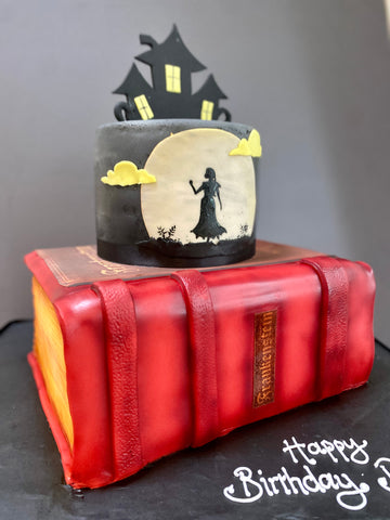 Halloween-themed birthday cake with a red book and woman silhouette against a big moon and a spooky house cake topper