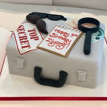 Birthday cake shaped like a briefcase with spy decorations on top like sunglasses, money, and a top secret sign