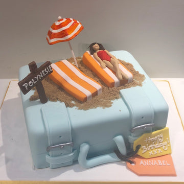 Cake designed like a suitcase with beach elements including an umbrella and lounge chair and a woman sunbathing