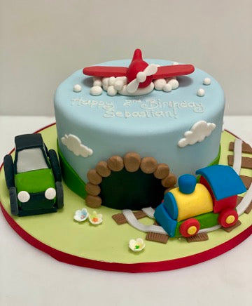 Children's birthday cake with train, car, and airplane decorations on a blue background.