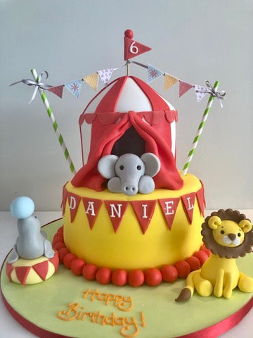 Birthday cake with circus theme featuring an elephant, lion, and red circus top tent.