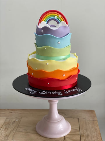two tiered birthday cake with a wavy rainbow around the sides and a sugar rainbow on top