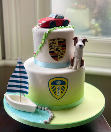 Two-tier cake with Porsche logo, toy car, and dog figurine on a table.