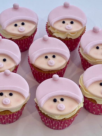Cupcakes decorated to resemble baby faces with pink hats and pink cheeks.