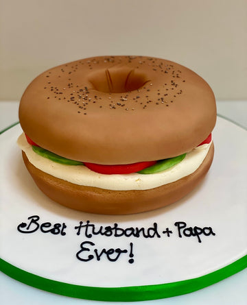 Cake designed like a bagel with cream cheese, tomato, and lettuce on a white plate with 'Best Husband + Papa Ever!' text.