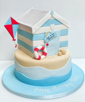 Cake designed like a beach scene with a striped beach hut, kite, and life buoy on a white background