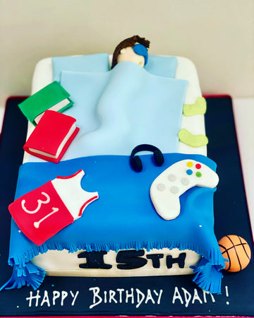 Birthday cake to look like a person in bed with a person figure, books, controller, and basketball on a blue blanket