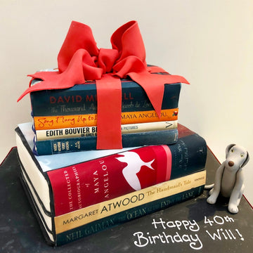 Cake decorated to look like a Stack of books tied with a red ribbon on a black surface, with a dog figurine a