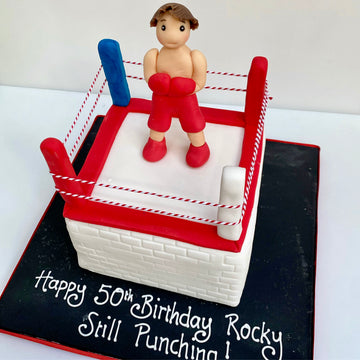 Cake decorated like a boxing ring with a figure of a boxer, on a white background.