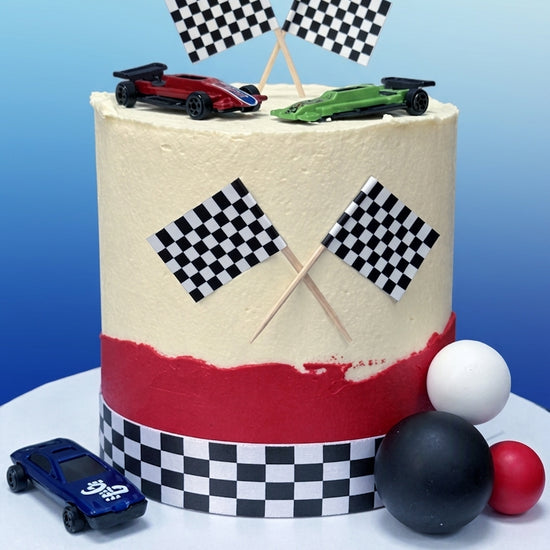A round frosted cake with a racing theme, featuring a cream top, a red band near the bottom, checkered flags, toy cars, and racing ball decorations on a white cake stand against a blue gradient background.