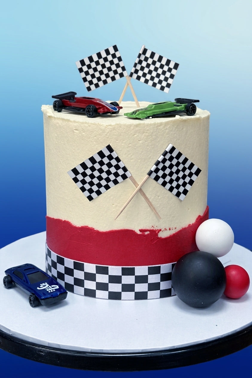 A round frosted cake with a racing theme, featuring a cream top, a red band near the bottom, checkered flags, toy cars, and racing ball decorations on a white cake stand against a blue gradient background.