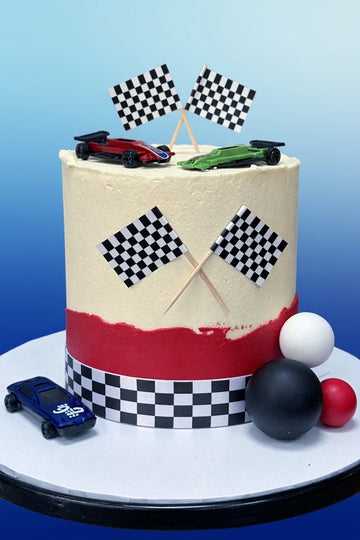 A round frosted cake with a racing theme, featuring a cream top, a red band near the bottom, checkered flags, toy cars, and racing ball decorations on a white cake stand against a blue gradient background.