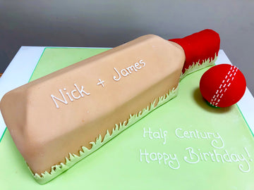 Cake shaped like a cricket bat with a red ball on a green base, featuring 'Nick + James' and 'Half Century Happy Birthday!' text.