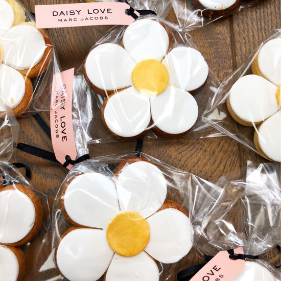 Daisy-shaped cookies with white icing petals and yellow centers, individually wrapped in clear cellophane bags with pink Marc Jacobs tags, arranged on a wooden surface.