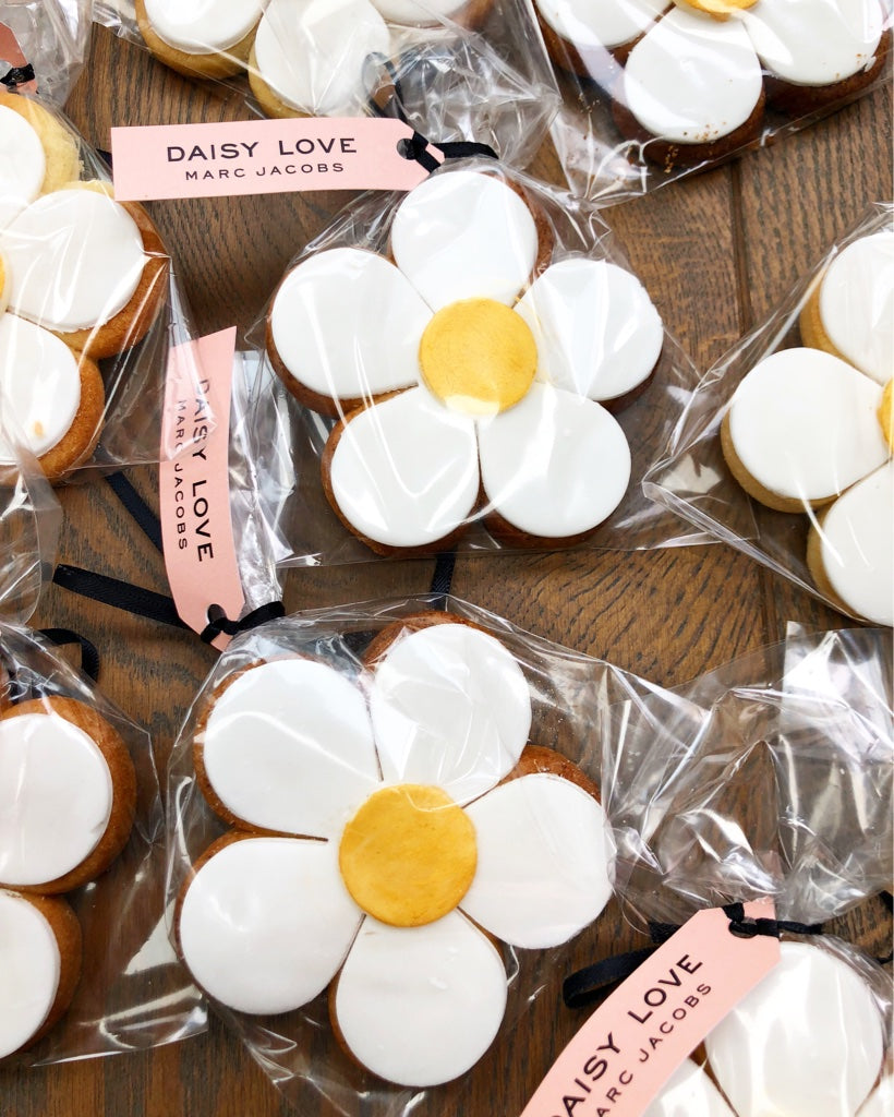 Daisy-shaped cookies with white icing petals and yellow centers, individually wrapped in clear cellophane bags with pink Marc Jacobs tags, arranged on a wooden surface.