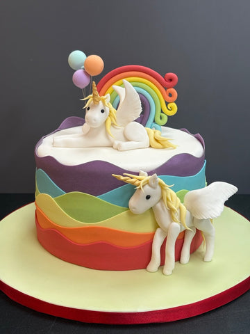 Wavy rainbow sides to a birthday cake with a curly rainbow on top and two flying unicorns