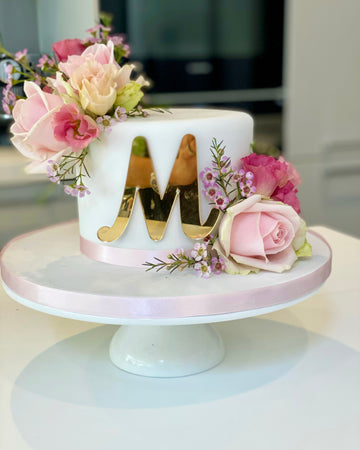 Beautiful Decorative cake with gold letter 'M' and pink flowers on a white stand.