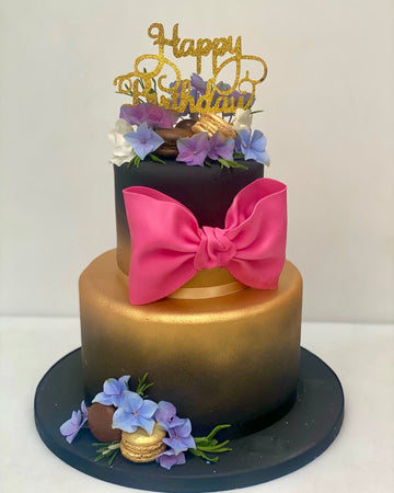 Two-tiered birthday cake with a pink bow, gold accents, and 'Happy Birthday' topper on a white background.