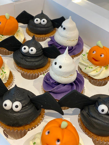 Halloween-themed cupcakes with bat, ghost, and pumpkin designs on a white surface.