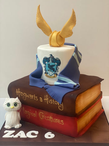 Large Harry Potter Birthday Cake with spell books as the bottom tier and the top tier with a gold snitch on top, a hogwarts scarf and harry potter's owl next to the cake