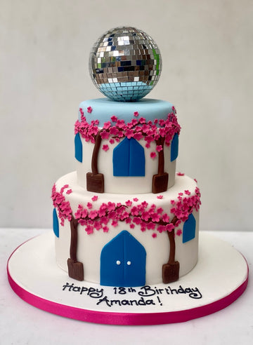 Two-tier birthday cake in the theme of mamma mia the musical with bougainvillea, blue doors, and a disco ball on top against a white background.
